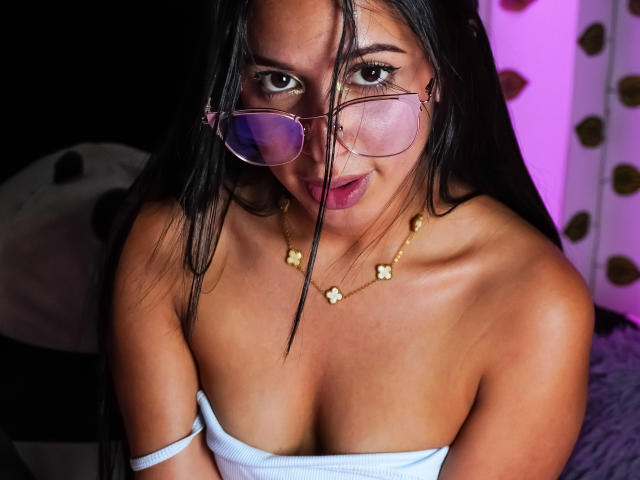 Ciamsth with long black hair, wearing pink glasses, a white strapless top, and a gold flower necklace, showing her tits. She has a cute, slightly open-mouthed, sexy expression.