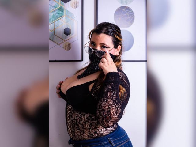 Salma Azaman mit Brille, schwarzes Spitzenoberteil, zeigt große Brüste, trägt eine schwarze Maske, hält die Maske mit der rechten Hand, blaue Jeans, steht an einer Wand, braunes Haar, weißer Hintergrund..