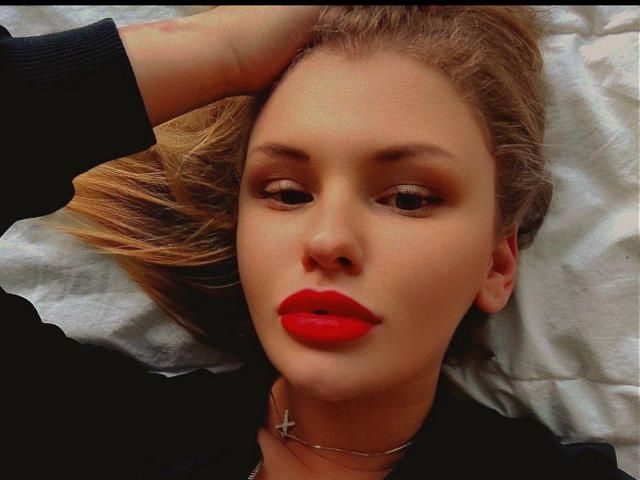 PassionFruitI has a cute face with red lipstick, lying on a bed with blonde hair, wearing a black shirt, and a silver cross necklace. Her hand's on her head.