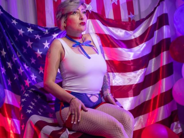 MaferCougar with short blonde hair, white tank top, blue ribbon, fishnet stockings, red choker, sitting on red chair, American flag, tattoos on right arm, purple and red lighting.