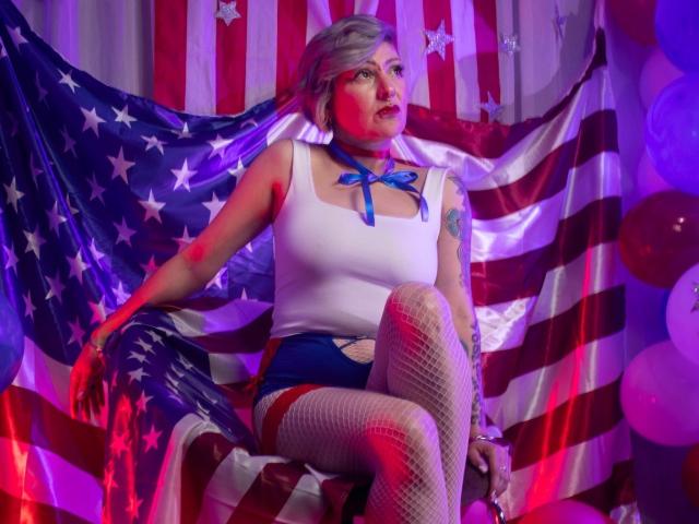 MaferCougar with silver hair, white tank, blue ribbon, fishnet legs, and red/white/blue shorts, sitting on chair, American flags behind, purple/blue/red lighting, looking off to side.