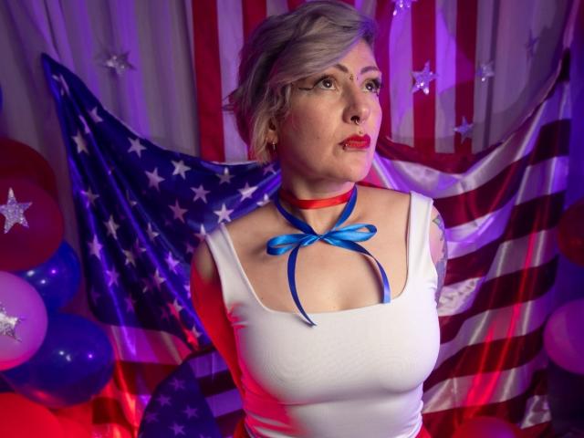MaferCougar with short blonde hair, red lipstick, white tank top, blue ribbon on chest, red choker, American flag backdrop, purple and red lights, purple balloons, looking slightly up.