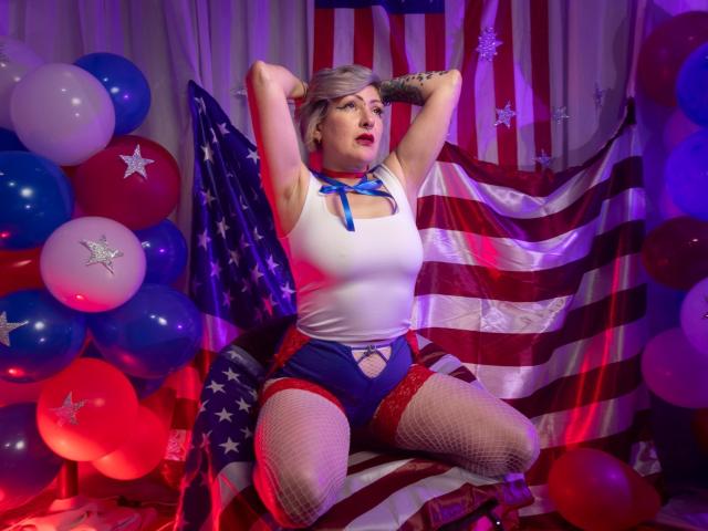 MaferCougar with gray hair, white tank top, blue ribbon, red shorts, fishnet stockings, and American flag background, posing with arms raised, red and blue balloons on the left.