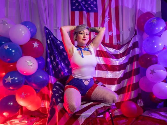 MaferCougar with short blonde hair, white tank top, blue shorts, fishnet stockings, and red ribbon, posing with arms behind head, American flag and balloons in colorful, festive setting.