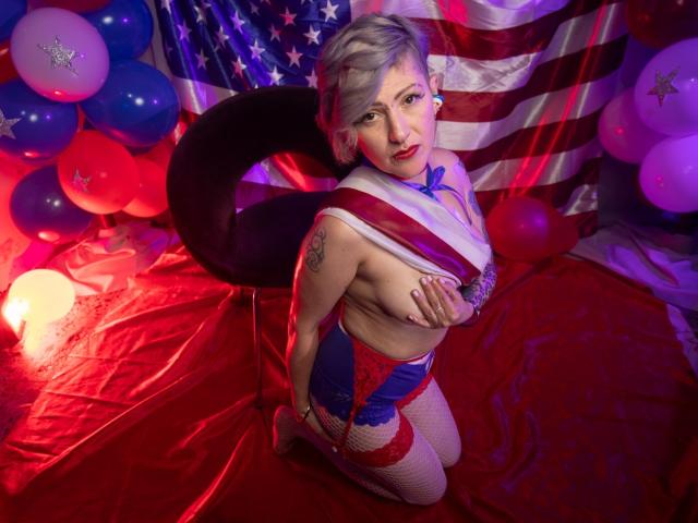 MaferCougar with short blonde hair, red and blue lingerie, fishnet stockings, covering her tits with one hand, kneeling on red fabric, American flag and balloons in the background.