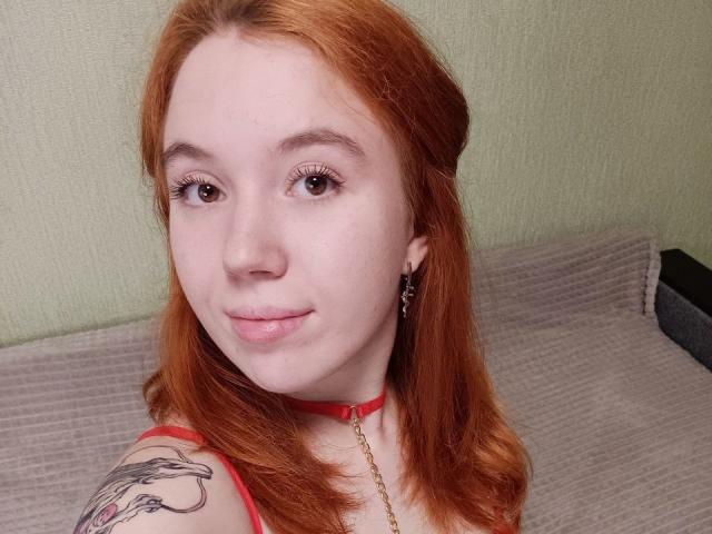 AnikaGlow with red hair, pale skin, and a tattoo on her shoulder, wearing a red choker, has shiny lips and a subtle smile, brown eyes, and eyeliner.