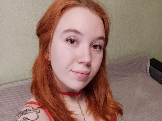 AnikaGlow has red hair, fair skin, and a small nose. She's wearing a red choker and has a tattoo on her left shoulder. Her eyelashes are long and brown.