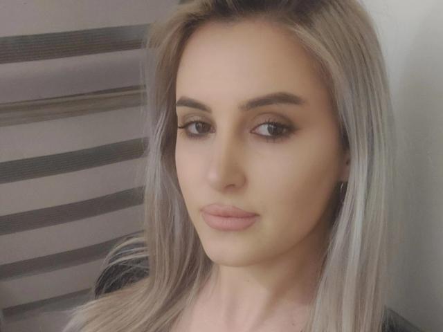 ElyAnna69 has blonde hair, brown eyes, and pink lipstick. She's looking slightly to the side with a neutral expression. Her makeup is subtle, and she's wearing a black top.