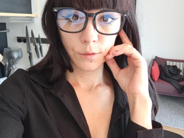 OrianaLaFrancaise with black glasses, reflection showing her blue eyes, wearing a black shirt, holding her chin, slight pimple on her cheek, brown hair, white skin.