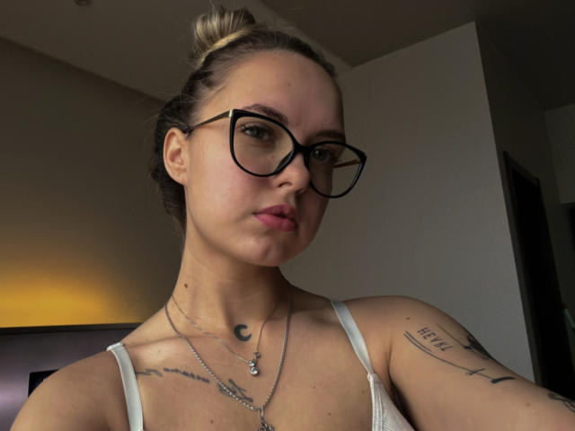 KhaleesiMoon tiene el cabello rubio recogido en un moño, gafas negras, labios rosados, una camiseta blanca, tatuajes en el cuello y el brazo, y un collar con forma de luna creciente, mirando a la cámara..