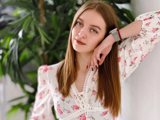 MaryJoy, avec ses longs cheveux bruns, portant une blouse fleurie, a un visage mignon, de petites seins, et une montre connectée à son poignet, et elle est appuyée contre une plante..