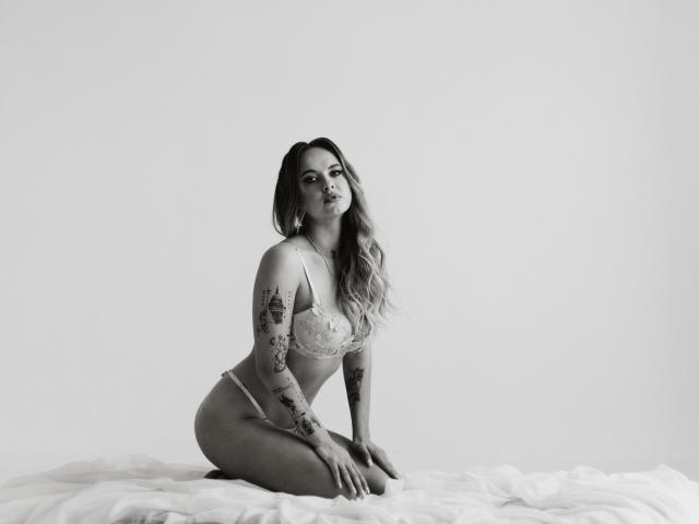 KhaleesiMoon, kneeling on white bed, wearing lace bra and panties, showing her tits and ass, tattoos on arms, long wavy hair, black-and-white photo, looking at camera.
