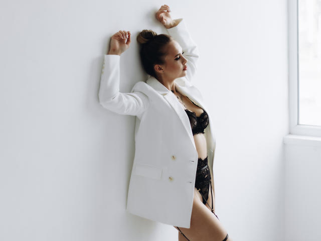 KhaleesiMoon, in a white jacket, leans against a wall, arms raised, wearing black lace lingerie with visible tits and pussy, in a brightly lit room.