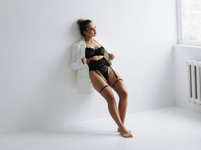 KhaleesiMoon in black lace lingerie with garter straps, open white jacket, standing against white wall, bare feet, looking to the side, light from window on the right.