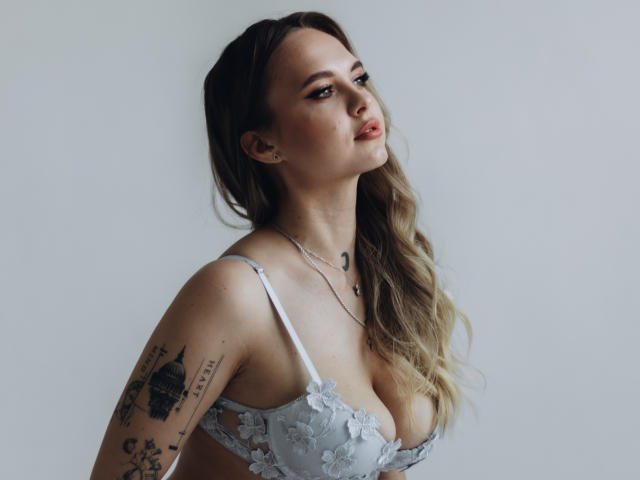 KhaleesiMoon with long, wavy blonde hair, wearing a light blue lace bra, showing her large tits and tattoos on her right arm, looking seductively at the camera.