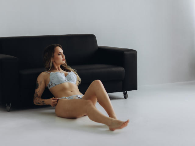 KhaleesiMoon with small tits and a slim ass, wearing light blue lace lingerie, sits on a white floor with legs extended, arms resting on a black sofa.