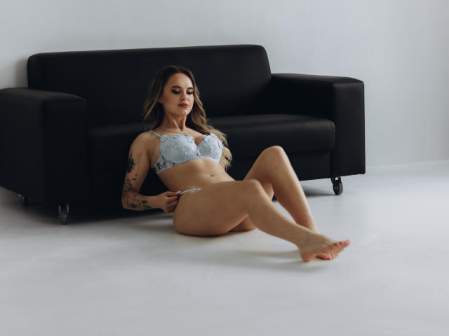 KhaleesiMoon with pale blue lace bra and matching panties, sitting on white floor, legs extended, black sofa behind, tattoos on right arm, wavy brown hair, soft expression.