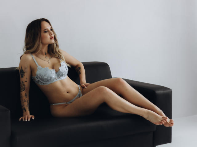 KhaleesiMoon with pale skin, blonde hair, tattoos, wearing light blue lace bra and panties, sitting on black couch, legs extended, looking to the side, sexy.