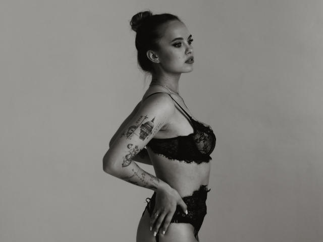 KhaleesiMoon with tattoos, wearing black lace bra and panties, side profile, hands on hips, dark hair in bun, looking forward, grayscale photo.