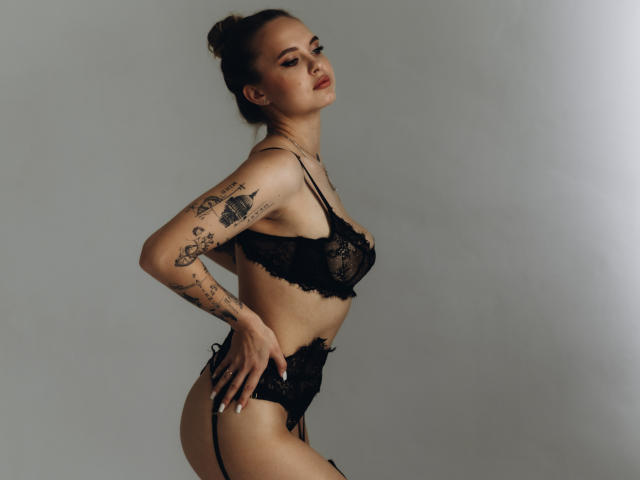 KhaleesiMoon in black lace lingerie, showing off her tattoos, with her hands on her hips, standing in profile, displaying her tits and ass. Her hair is in a bun.