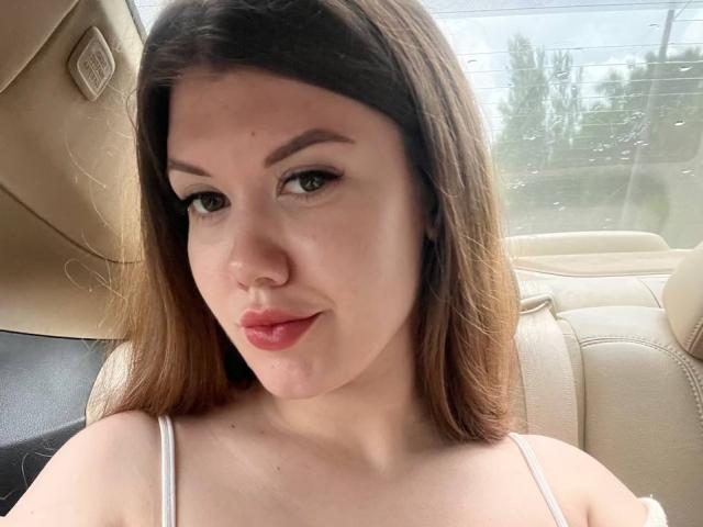 Nikki, con la pelle chiara, gli occhi marroni e il rossetto rosso, ha i capelli castani lisci e indossa una canotta bianca a spalline, seduta in un'auto beige..