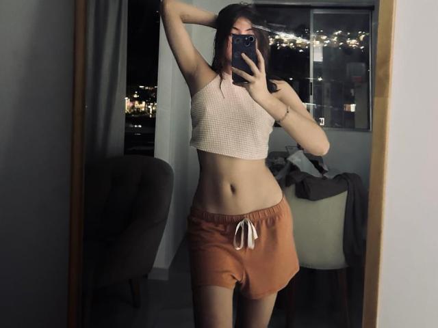 SpencerLe, con un fisico snello e piatto, indossa un top corto rosa e pantaloncini arancioni, tiene un telefono in un selfie allo specchio, con le braccia alzate, mostrando il seno e i glutei..