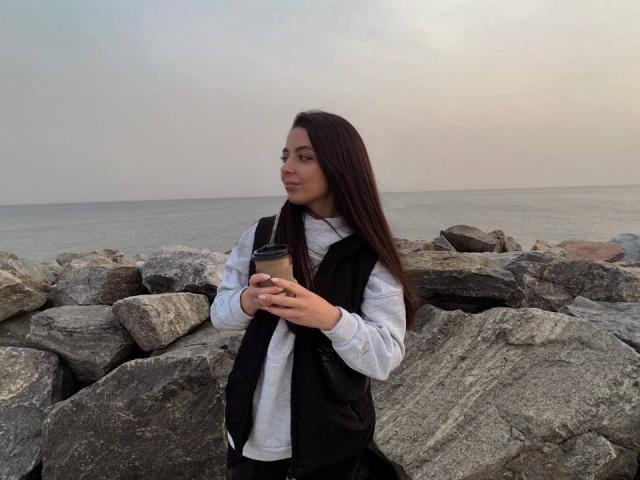 DiamondEyesss with long dark hair, wearing a black vest over a gray shirt, holding a coffee cup, standing in front of large rocks by a gray, calm ocean.