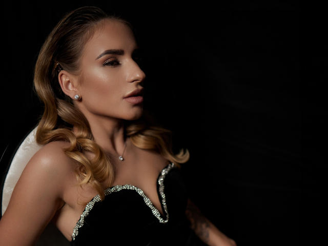 LaymaBlast with blonde curls, wearing a black dress with silver trim, has shiny skin and a necklace, gazing sideways with a serious expression, against a dark background.