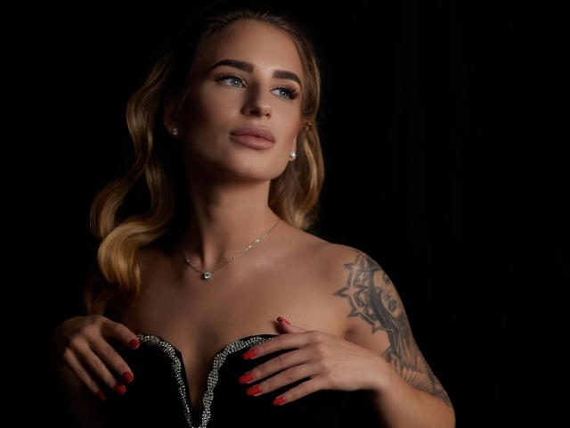 LaymaBlast with blonde hair, blue eyes, and a tattoo on her right arm, wearing a black, strapless top with a silver zipper, has red nails and a silver necklace.