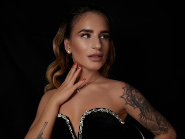 LaymaBlast, con coletas rubias, vestida con un vestido negro sin tirantes con detalles plateados, tiene tatuajes en su brazo derecho, esmalte de uñas rojo y un arete de diamantes, mirando hacia un lado..