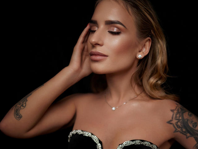 LaymaBlast con le mani sul viso, indossa un top scintillante senza spalline, orecchini di diamanti, tatuaggi sulle braccia, trucco con glitter, capelli castani ondulati, sfondo nero, espressione sicura, sexy..