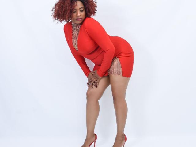 LiloTorontoo has curly red hair, a red dress with a deep cleavage, and red high heels, bending forward with hands on knees, showing her ass and a peek of her pussy.