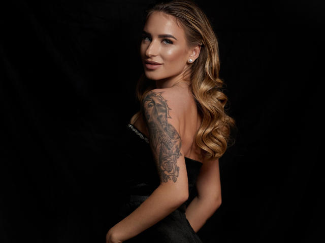 LaymaBlast com cabelos loiros longos, vestido preto sem alças, olhando para trás, mostrando o braço tatuado, sorriso discreto, fundo escuro, brincos, sexy, atraente..