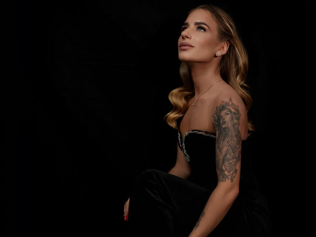 LaymaBlast, com cabelo loiro ondulado, vestido preto sem alças, braço tatuado e brincos, olhando para cima, sentada contra um fundo escuro. Seus seios são visíveis sob o vestido..