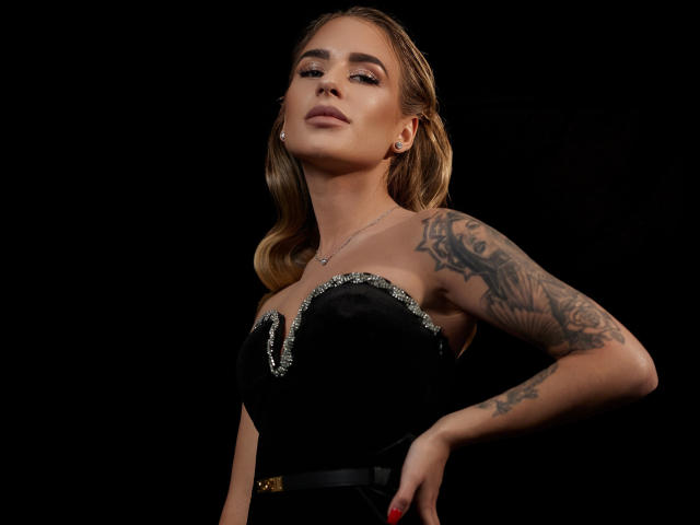 LaymaBlast com cabelo loiro ondulado, vestido preto sem alças, blusa brilhante, braço tatuado, pose confiante, fundo preto, maquiagem discreta, esmalte vermelho, brincos..