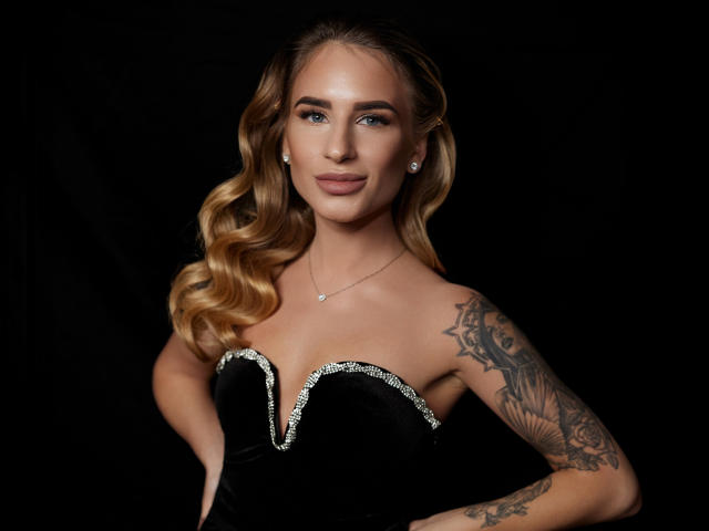 LaymaBlast tem cabelo loiro ondulado, olhos azuis e uma tatuagem no braço direito. Ela está vestindo um vestido preto sem alças com detalhes prateados e um colar delicado..