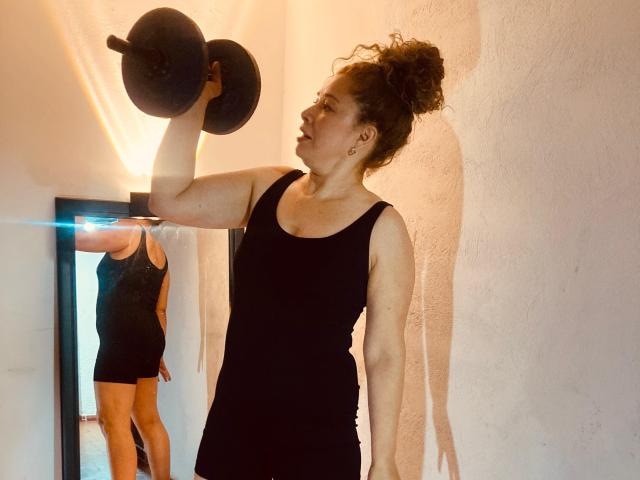 DalilaSuperHot with curly hair in a black tank top and shorts, lifting a dumbbell, reflected in a mirror, showing her toned arms and ass, in a sunlit room.