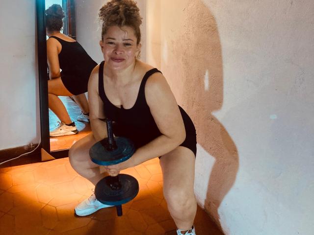 DalilaSuperHot with curly hair, black tank top, and white sneakers, squatting with a dumbbell, showing her ass in a mirror reflection, smiling, shadow on the wall.