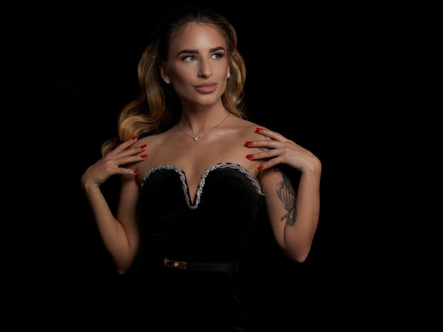 LaymaBlast with blonde pigtails, wearing a black strapless dress with silver trim, red nails, hands on chest, tattoo on right arm, black background, looking to the side.