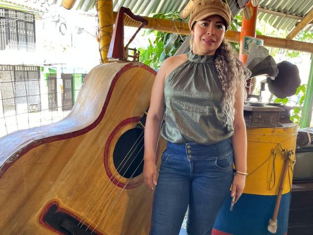 DalilaSuperHot stands with a curvy ass in blue jeans, shiny silver top, and brown cap, next to a giant wooden guitar. Long curly hair, smiling, outside.