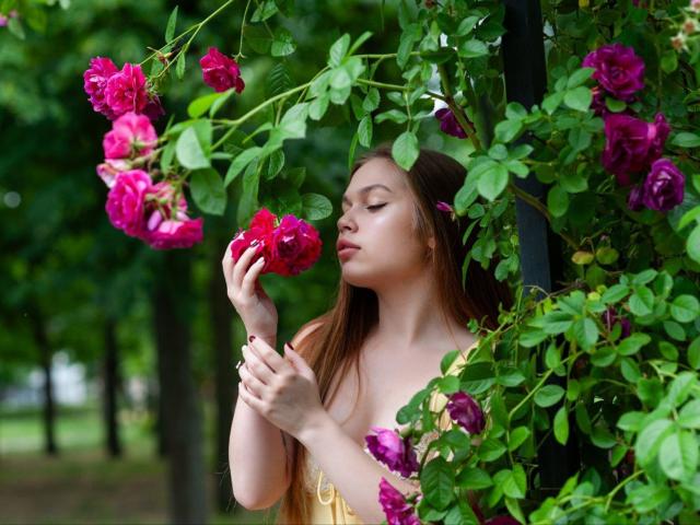 LaraPika with long brown hair, eyes closed, holding a pink rose to her nose, surrounded by vibrant pink and purple roses, green leaves, and a black pole.