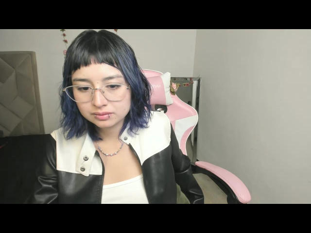 Aiiko with blue-dyed hair, glasses, and a black-and-white leather jacket, sitting on a pink gaming chair, pouting with her lips slightly curled.