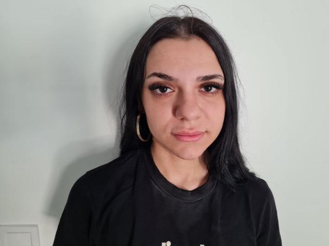 MiniMira has dark, straight hair, brown eyes, and wears a black shirt with gold hoop earrings. Her face is slightly flushed, and she's looking at the camera with a slight smile.