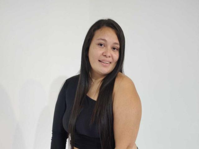 InaraBeki with medium tits, black long-sleeve dress, smiling, dark straight hair, light brown skin, standing against white wall.