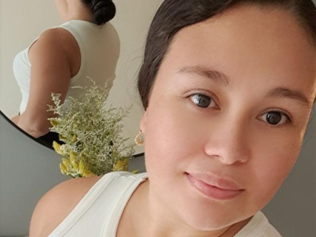 InaraBeki has light brown skin, black hair in a bun, wearing a white tank top, showing a subtle smile, with small gold earrings, and a reflection of her back.