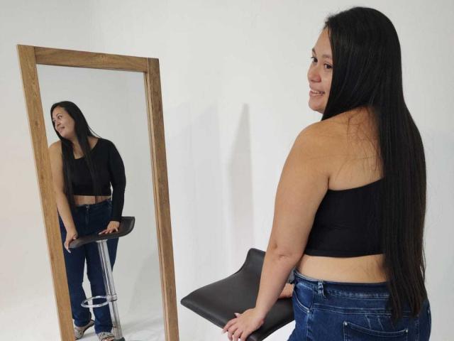 InaraBeki with long black hair, wearing a black crop top and blue jeans, stands by a mirror, showing her back and smiling at her reflection. She's leaning on a stool.