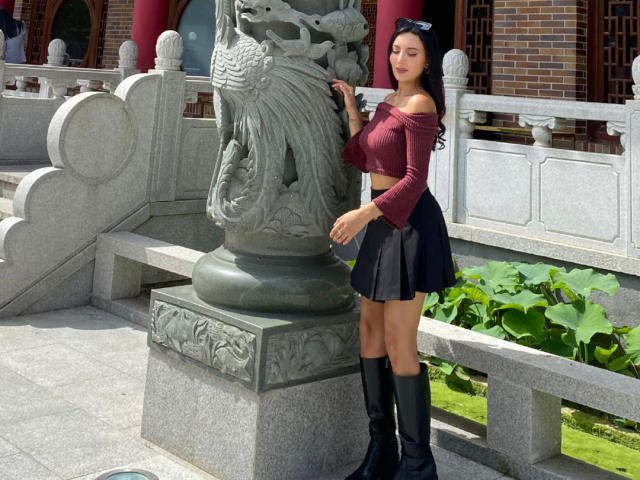 CarlaFranco with long black hair, wearing a red off-shoulder top, black skirt, and black knee-high boots, standing next to a green dragon statue, holding its base.