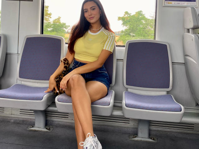 CarlaFranco, com longos cabelos castanhos, está sentada em um assento de trem, vestindo uma camiseta amarela, shorts escuros, tênis brancos e uma pulseira com estampa de leopardo, exibindo suas pernas e nádegas tonificadas..