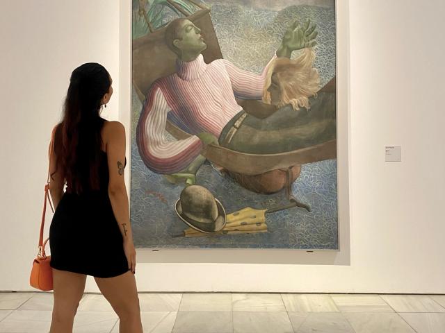 CarlaFranco stands with her back to the camera, wearing a black dress and orange bag, gazing at a painting of a green-skinned man in a striped shirt.