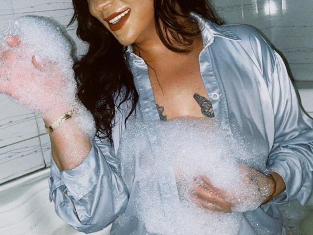 LorenaLoveS with wet, wavy brown hair, smiling, wearing a shiny blue shirt, covered in soap bubbles, showing small tits, tattoo under shirt, right hand raising bubbles.